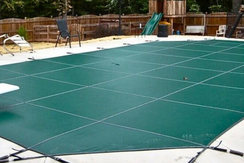 Green safety mesh winter pool cover installed over an inground pool with diving board and patio deck.